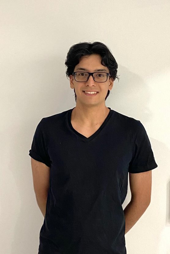 An individual with short wavy hair with glasses, in a black t-shirt smiling at the camera, against the backdrop of a white wall