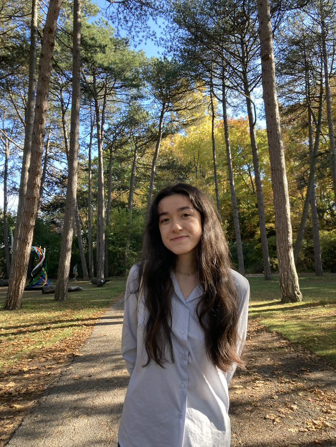 An individual with long dark hair, wearing a light blue shirt, smiling at the camera, against the backdrop of nature.