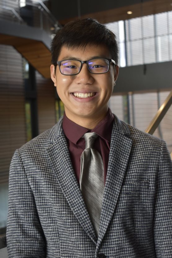 An individual with short hair and glasses in a suit, smiling, against the backdrop of a large indoor space