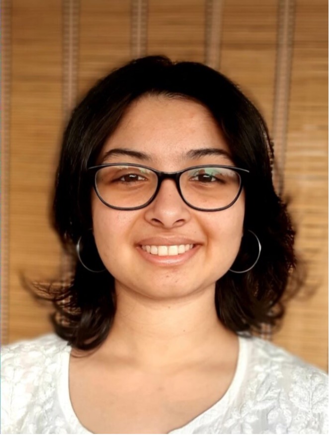 shoulder up image of Anoushka Indira Kapoor, a woman wearing black glasses with shoulder length hair and a white shirt, smiling