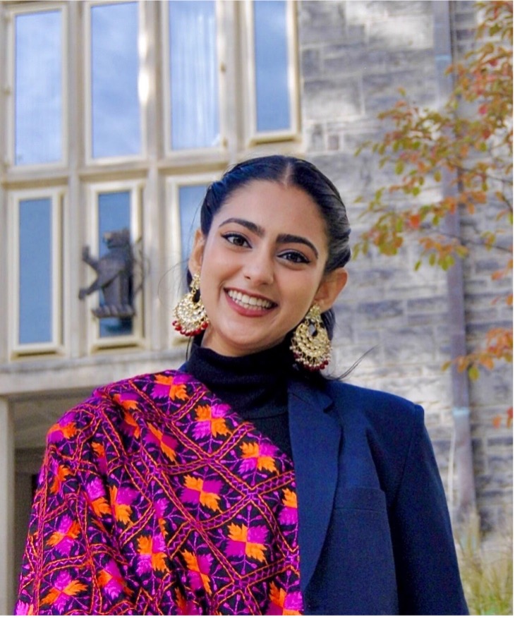 A photo of Bhavnoor Kaur Pannu from the waist up, a woman standing outdoors wearing a pink patterned sari with a blazer, smiling.