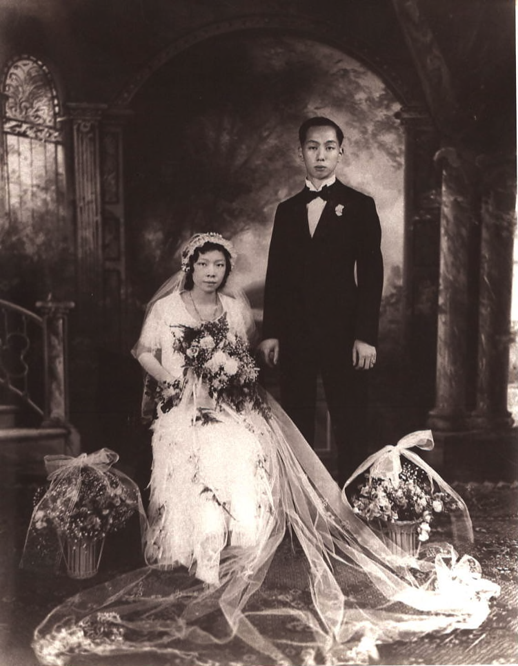 A Chinese couple on their wedding day in the 1930s. The bride is wearing a long white dress and the couple are flanked by two potted flowers.