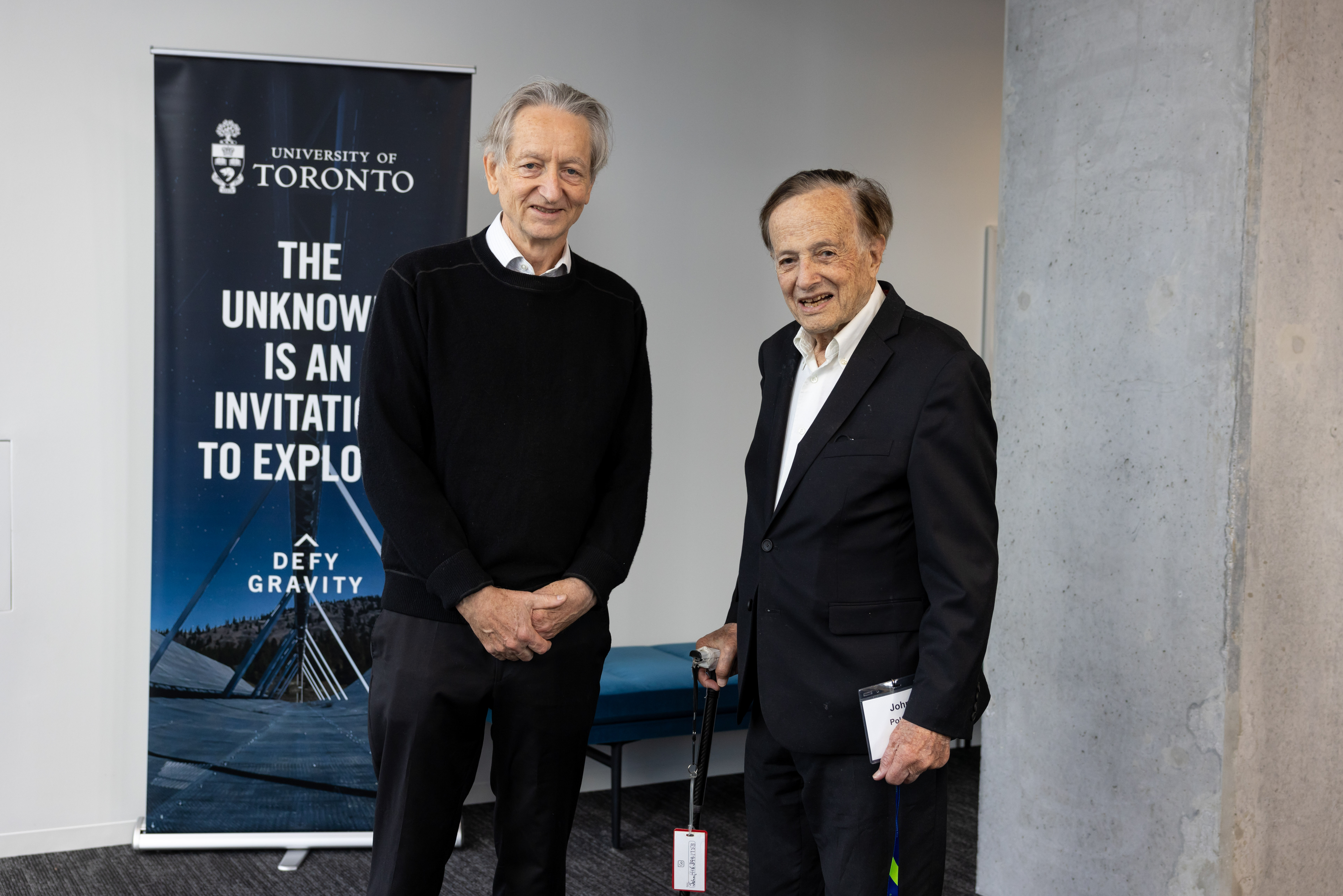 Two white men in their 60s and 70s stand in front of a grey wall. One of the men is holding a cane.