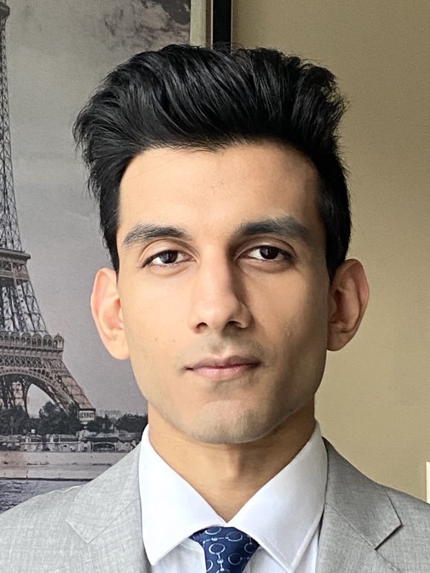 An individual with short dark hair, wearing a suit, against the backdrop of a a painting of the Eiffel tower.