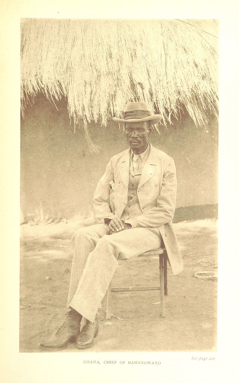 An image of Khama III, a Black Botswanan leader in a tan suit, boots and a fedora in the 1930s. he is seated with his hands folded on his lap.