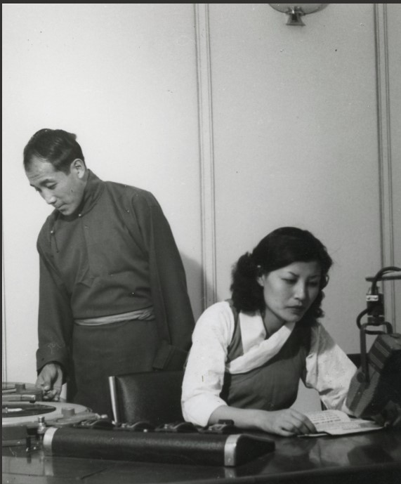 A Tibetan man in a traditional high necked buttonedshirt and his wife, a dark haired Tibetan woman in a pinafore and white blouse, are preparing to read for a live radio broadcast, with the woman bent over a script in front of a microphone.