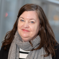 headshot of Loryl Mcdonald, a caucasian woman with brown shoulder length hair wearing a grey scarf, black sweater, and black and white striped shirt.