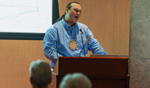 Drummer Daniel Secord, an Indigenous man in a blue shirt wearing a beaded medallion, performs drumming and singing before the audience at the Chamberlin Lecture on October 22nd, 2024.