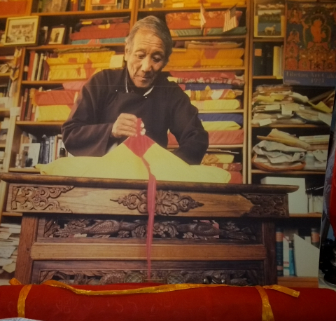 A Tibetan man with grey hair opens a traditional Tibetan text wrapped in beige cloth with red tie. A stack of colourful texts is piled behind him.